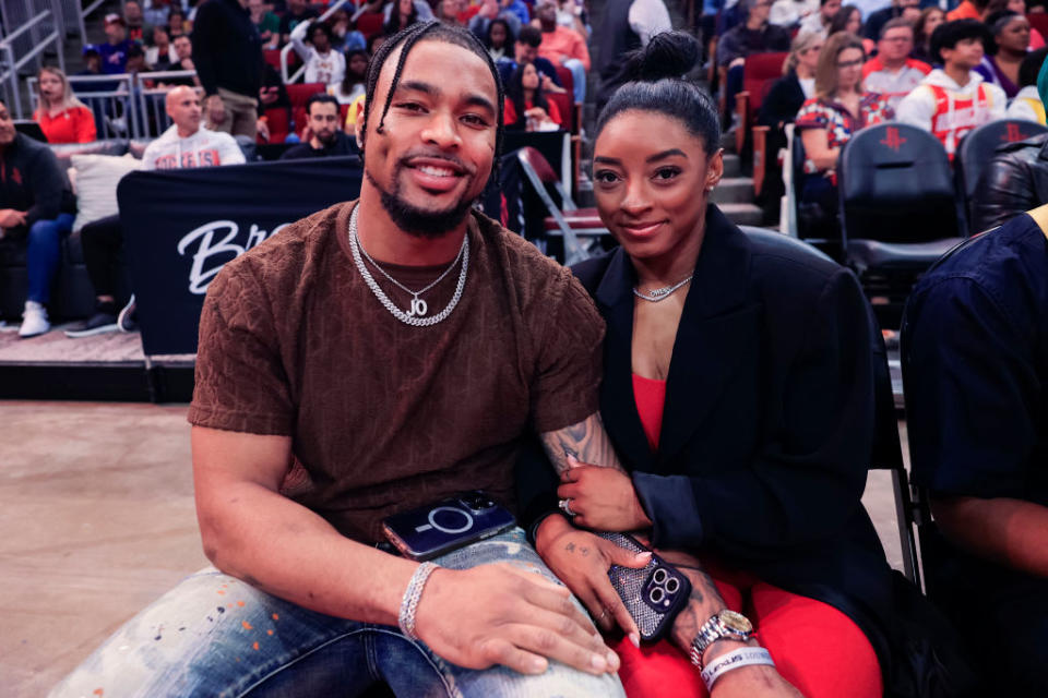 Jonathan Owens and Simone Biles smiling and holding hands while seated at a basketball game