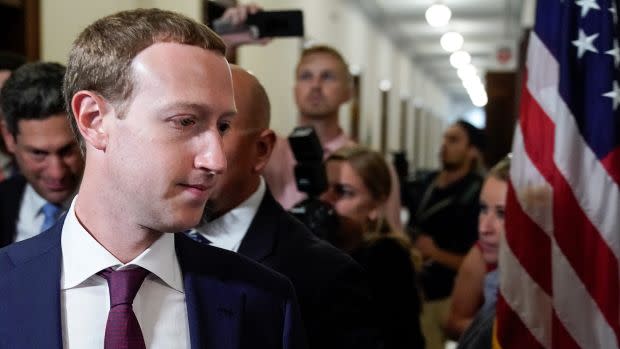 Facebook CEO Mark Zuckerberg looks glumly off the the side while surrounded by members of the press and an American flag.