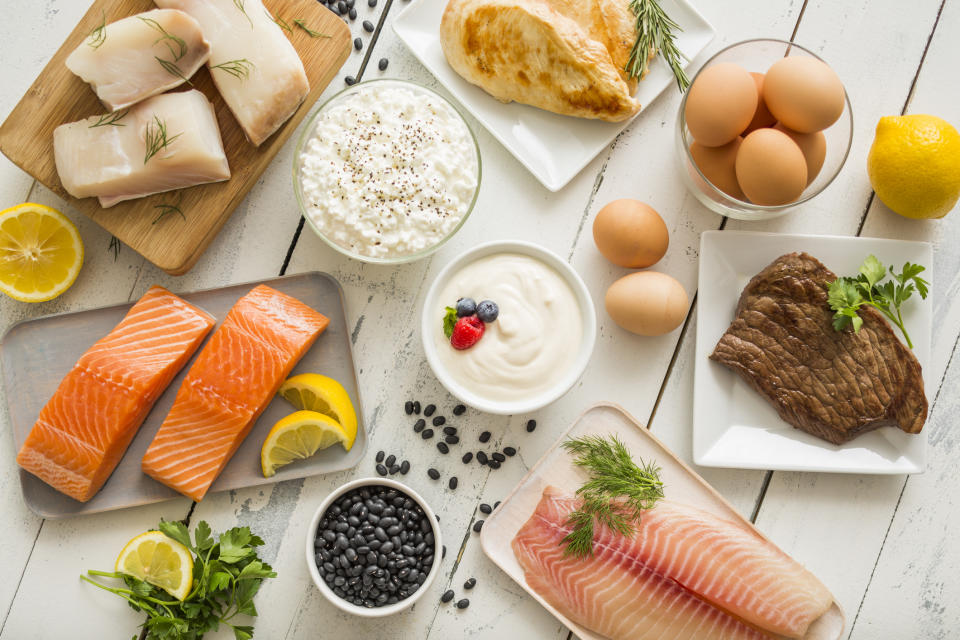 Various protein-rich foods displayed, including salmon, chicken, eggs, beef, cottage cheese, and yogurt, on a wooden surface with lemons and herbs