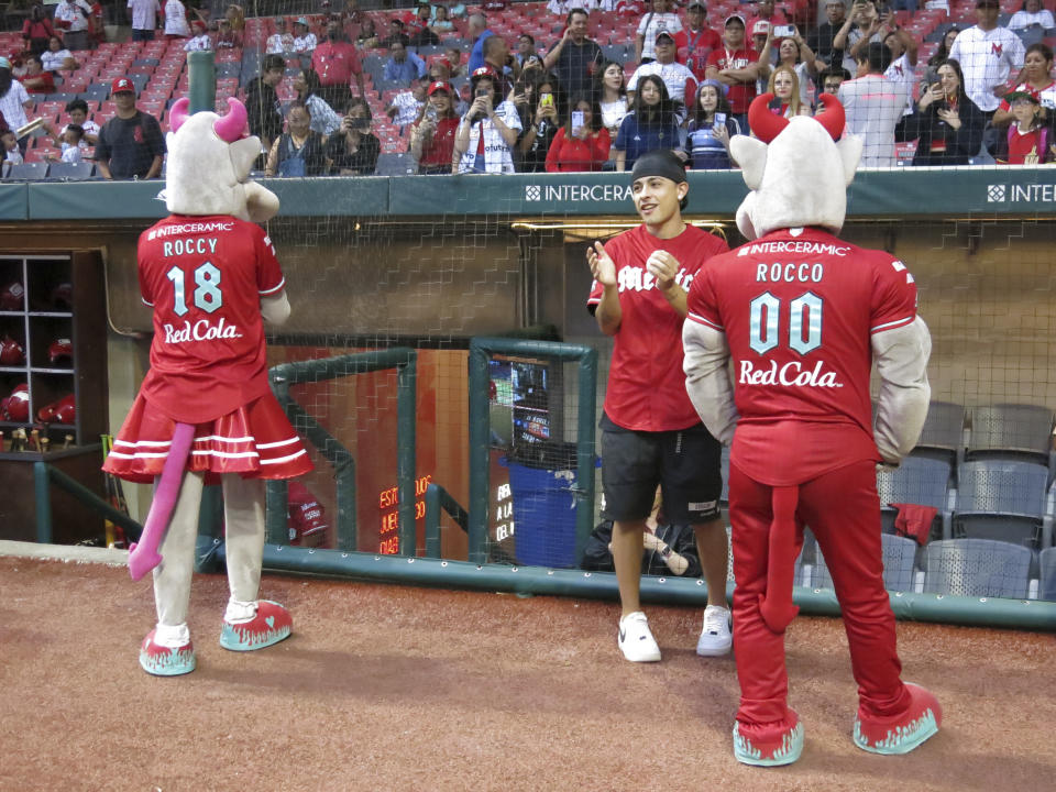 El rapero argentino Trueno, centro, se prepara para lanzar la primera bola del partido entre los Diablos Rojos del México y el Águila de Veracruz junto a las mascotas de los Diablos Rojos Roccy, izquierda, y Rocco en la Ciudad de México el jueves 2 de mayo de 2024. (Foto AP/Berenice Bautista)