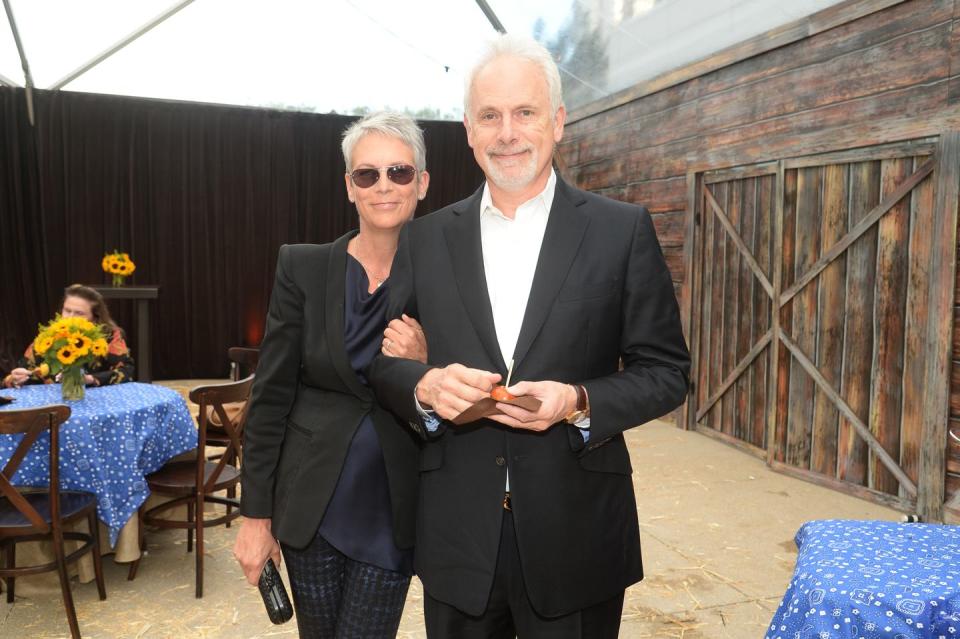 jamie lee curtis and christopher guest smile for a photo while standing together, they both wear black blazers and hold items in their hands