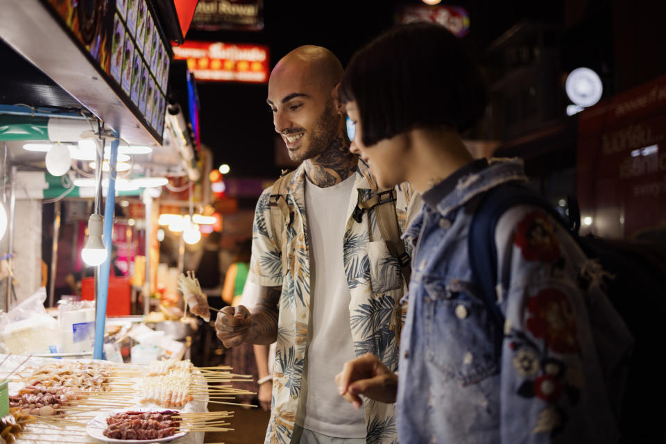 A man and a woman look at food skewers at a night market food stall. The man has tattoos and wears a floral shirt; the woman wears a denim jacket