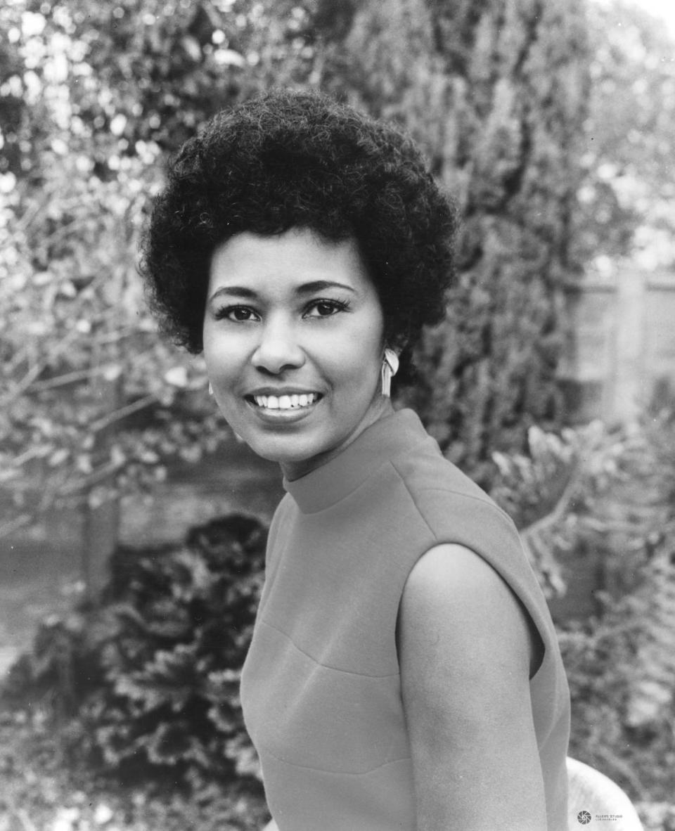 yvonne brathwaite burke smiles at the camera while sitting in profile and looking over one shoulder, she wears a sleeveless top and earrings, greenery is in the background