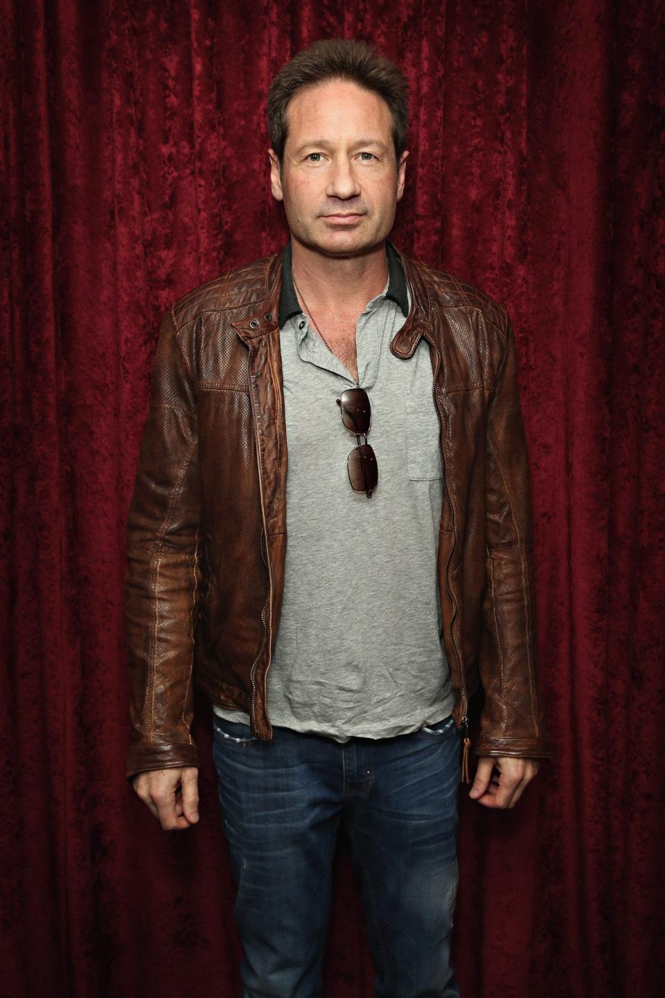David Duchovny, wearing a brown leather jacker and gray polo, smiles in front of a red curtain