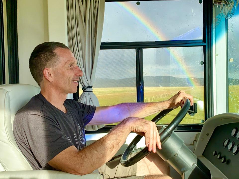 A man drives an RV with a rainbow in the background.