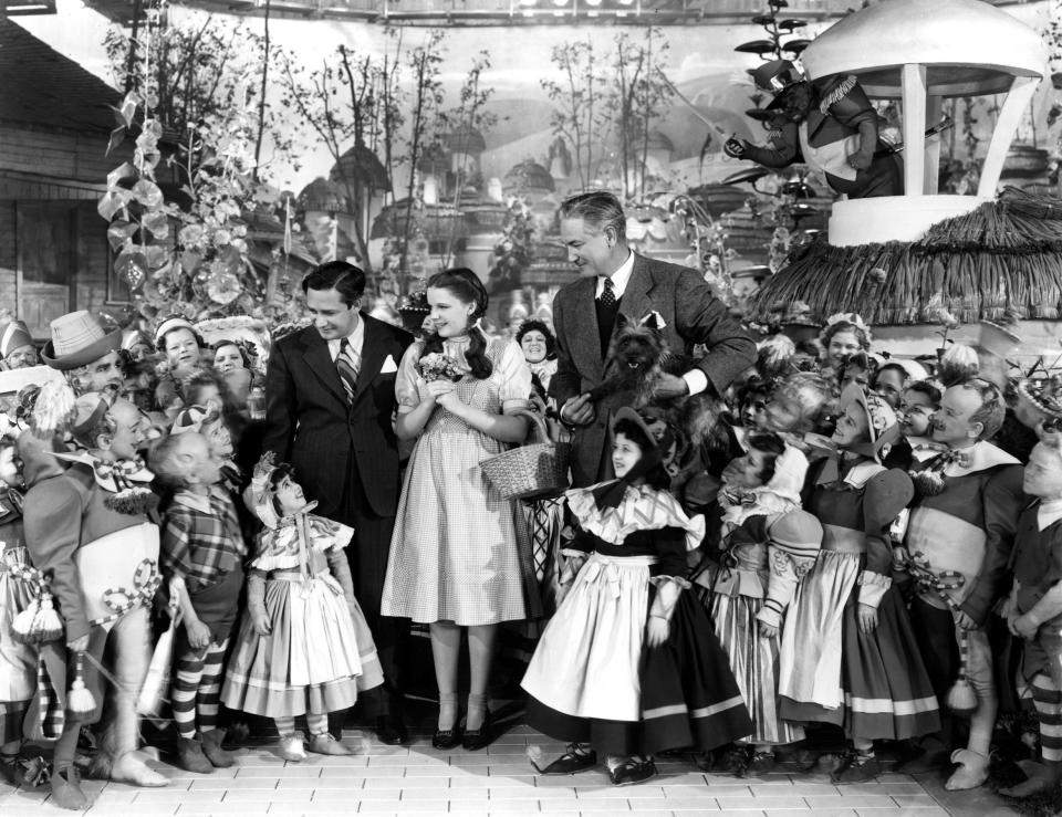 ”The Wizard of Oz’ producer Mervyn LeRoy, star Judy Garland and director Victor Fleming pose with Munchkin actors and Toto on the set in 1939 (Everett Collection)