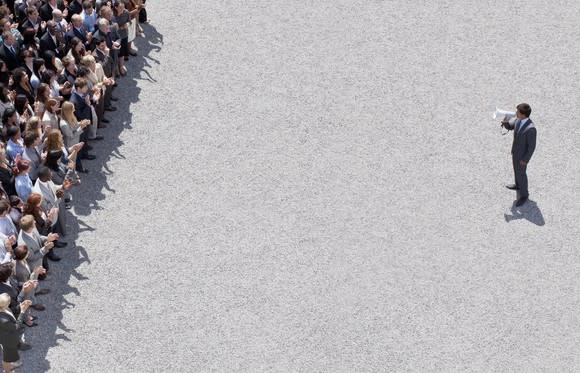 Man with a megaphone separated from a large crowd by a wide stretch of bare concrete.