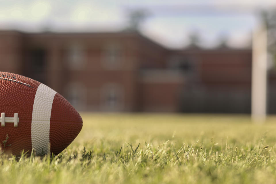 Football on grass stadium on college or high school campus. Goal post, school in background. No people.  Daytime.