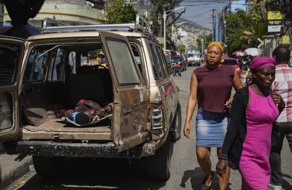 People walk past a forensics van holding a bloodied body after the body was removed from the sidewalk in Port-au-Prince, Haiti, Wednesday, May 1, 2024. Forensics said the body had gunshot wounds. (AP Photo/Ramon Espinosa)