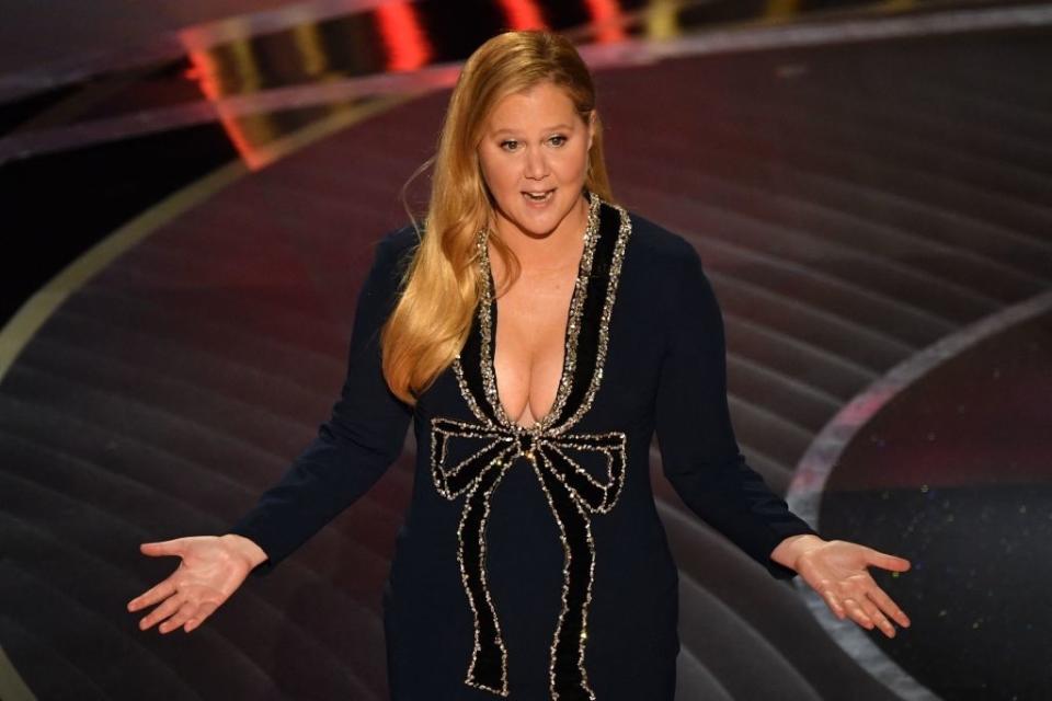 Amy Schumer on stage, wearing a deep V-neck gown with sparkling embellishments along the neckline and across the front, resembling a large bow