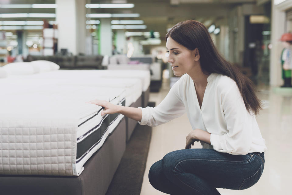 A woman in a white shirt and jeans in a mattress store. She examines the mattress she wants to buy. She squats and looks at the mattress