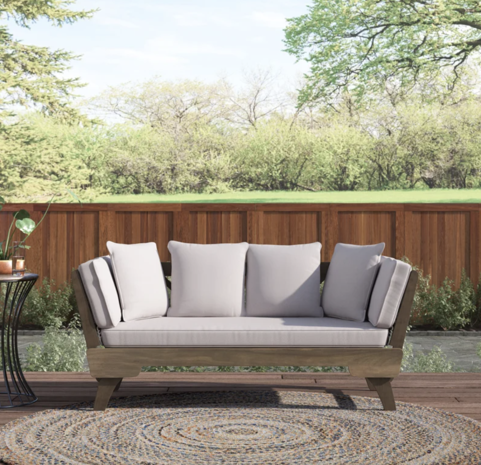 Roni Outdoor Patio Daybed with Cushions outside against wood fence and green trees (Photo via Wayfair)