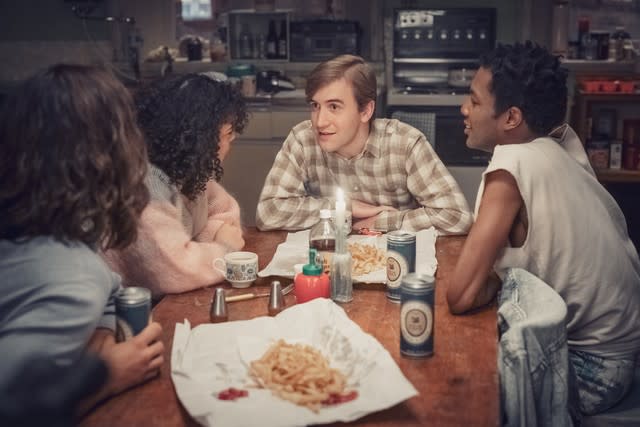 A group of people sit at a table talking, eating and drinking in a scene from "It's a Sin."