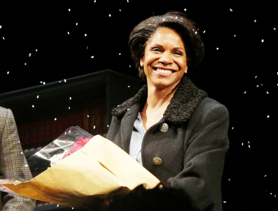 audra mcdonald wearing a black coat and blue shirt, holding flowers and smiling on a stage with a dark backdrop