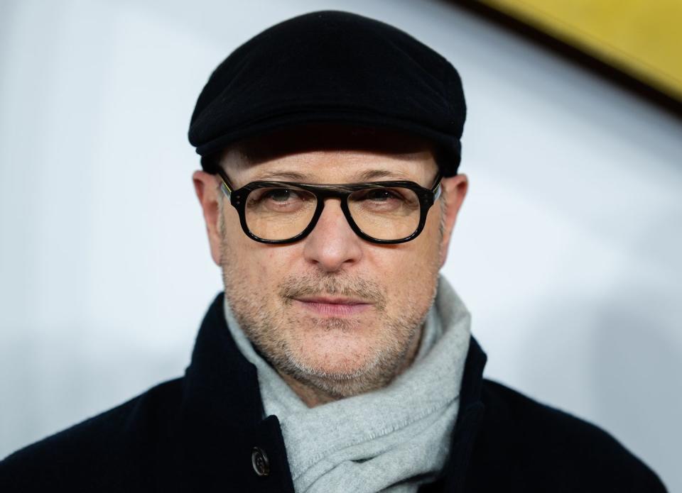 a closeup of director matthew vaughn wearing a pair of thick framed glasses, a black flat cap, a light gray scarf and a black coat