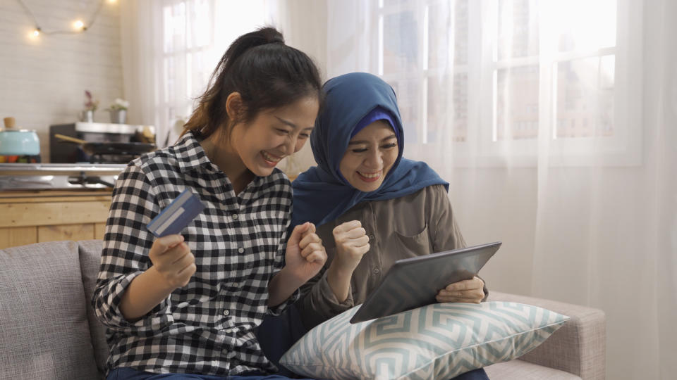 Cheerful multi female best friends triumphing while shopping online with digital tablet and credit card. two asian malay girls doing payment on internet by debit card relax sit on couch at cozy home