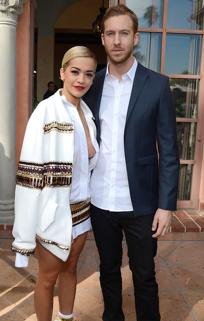 Rita Ora and Calvin Harris pose together; Rita wears an oversized outfit with detailed embroidery, and Calvin is in a button up shirt and dark blazer
