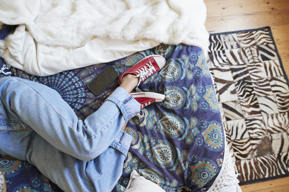 Person in blue jeans and red sneakers reclining on a patterned bedspread with a smartphone nearby. Image taken from above