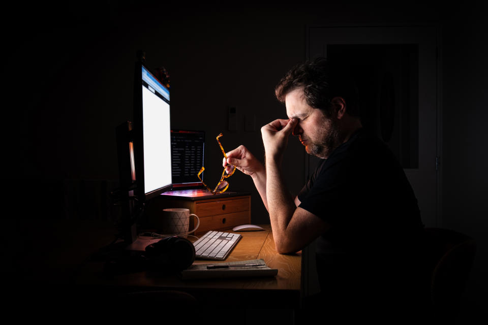 A man sits at a desk, rubbing his eyes while holding glasses in one hand, in front of a computer monitor and keyboard late at night