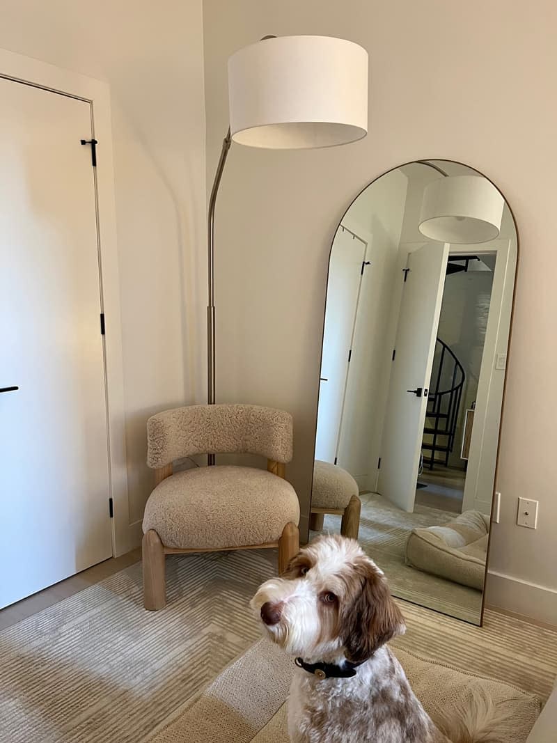 Drum shade floor lamp arcs over beige chair in corner next to leaning mirror near entrance.