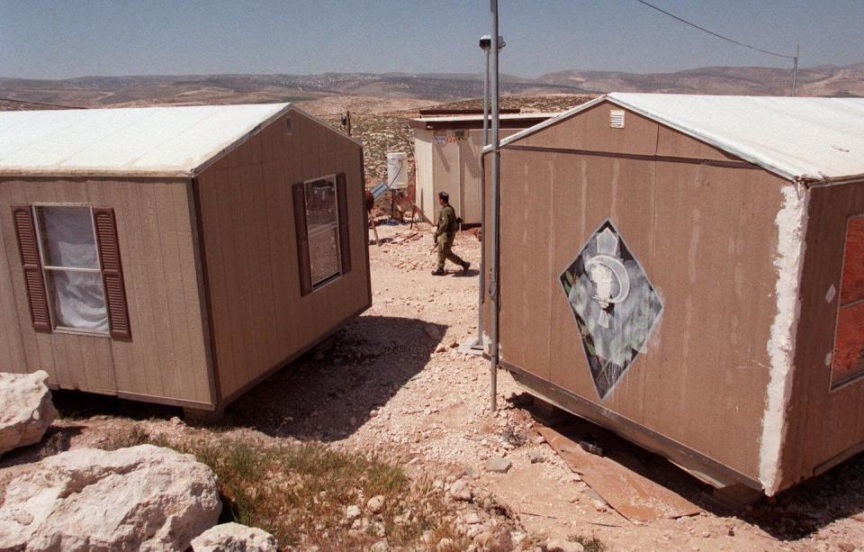 A person in an olive uniform walks between two tan-colored mobile homes on a hill in a dry landscape.