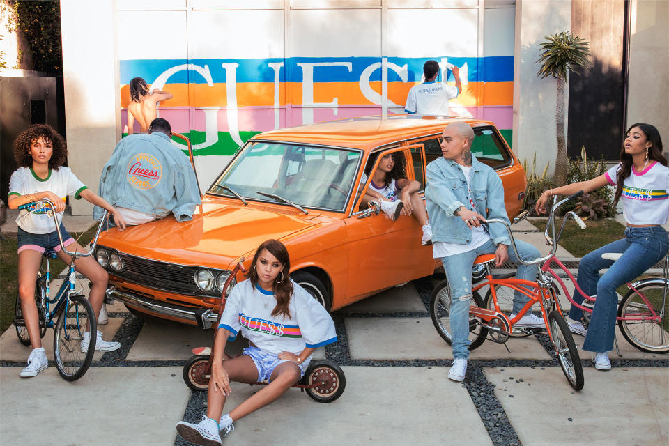 Several models wearing jeans and jean shorts in front of an old orange car in front of a Guess logo on the back wall.