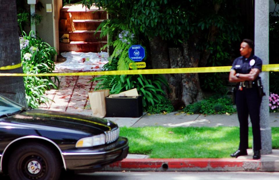 a view of a sidewalk and steps that are tapped off with yellow caution tape, blood stains and a white sheet are near the stairs, a police office stands on the curb and looks left as a car sits on the street on the left