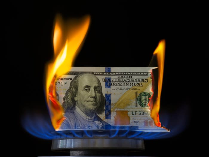 A one hundred dollar bill on fire atop a lit stove burner.