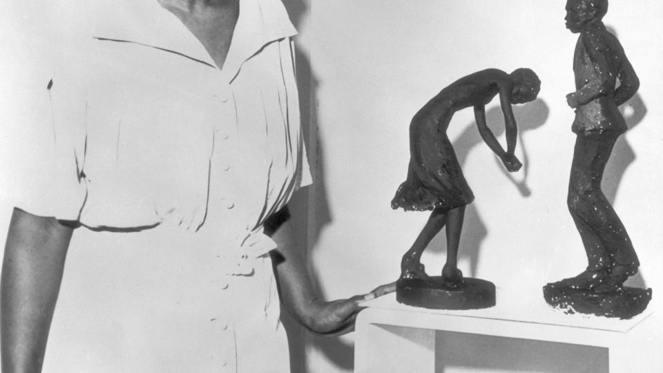 augusta savage resting her left hand on a table as she looks at two of her sculptures