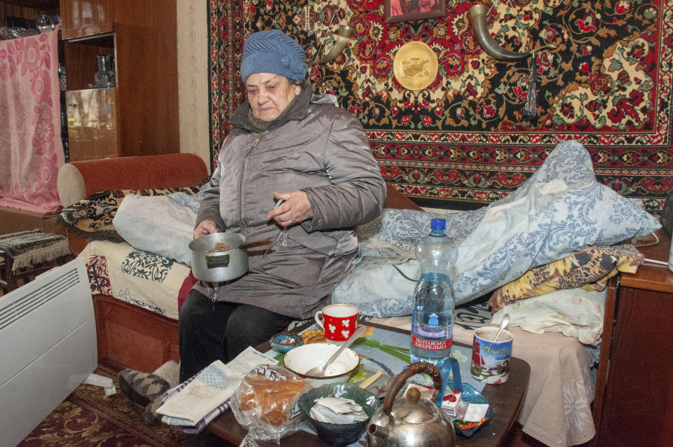 An elderly woman has a meal in her apartment, in Kharkiv, Ukraine, Sunday, March 13, 2022. The woman wears her coat and hat to keep warm as the municipal heating in the apartment block stopped after shelling. (AP Photo/Andrew Marienko)