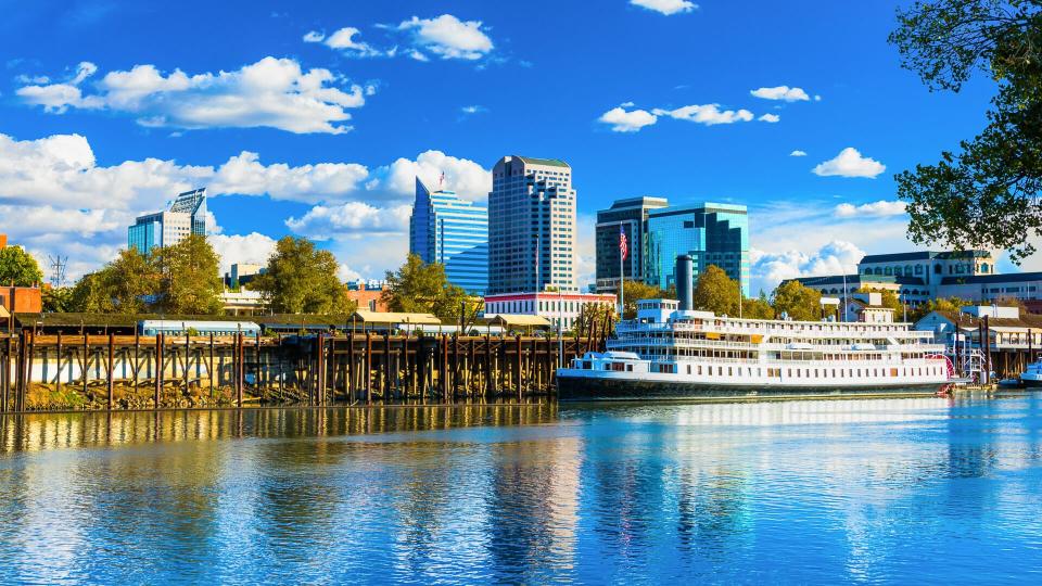 Downtown Sacramento skyline with the Sacramento River and the historic Delta King riverboat in the foreground and puffy white clouds and a deep blue sky in the background.