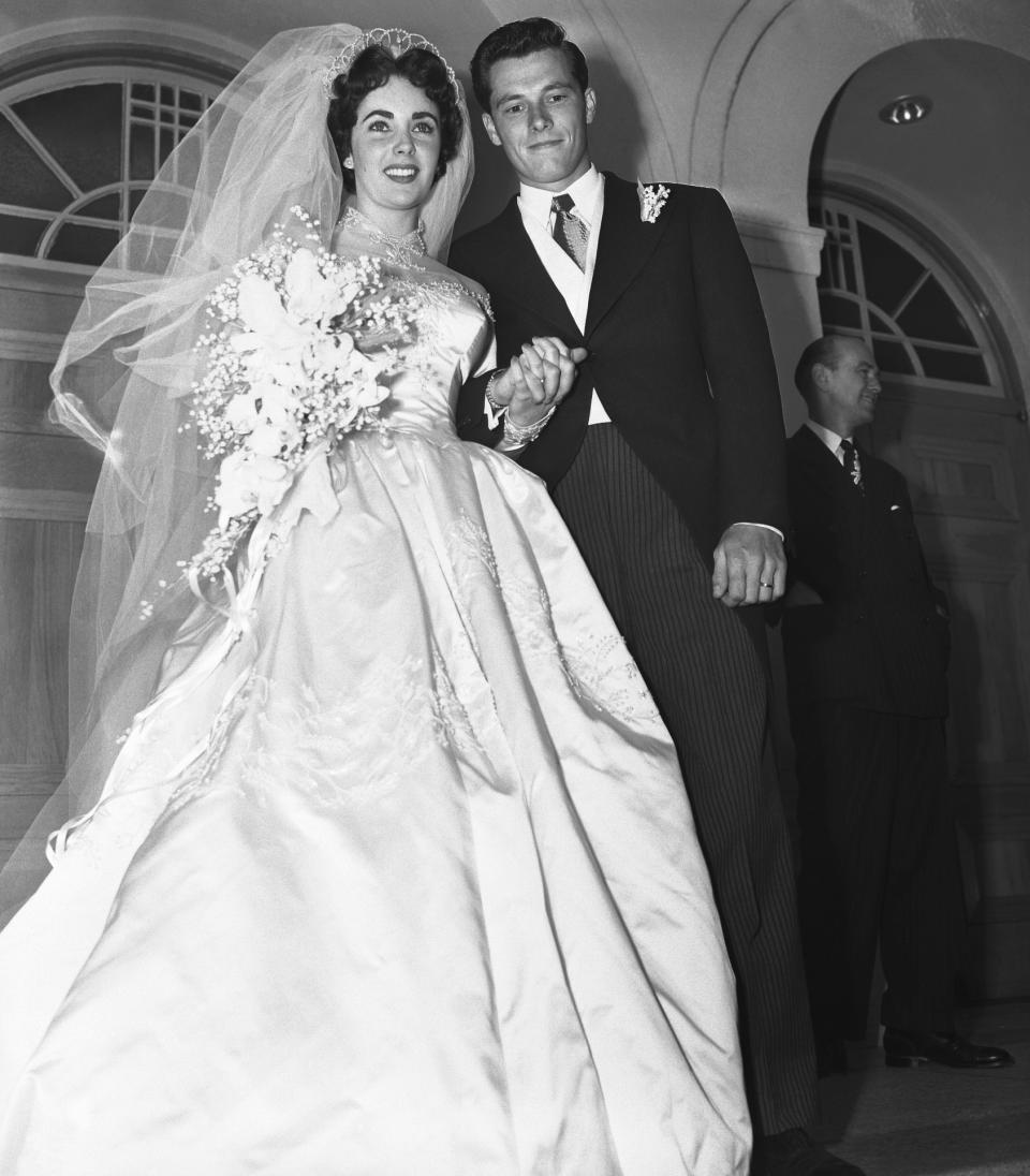 Newlyweds, the bride in a gown with a voluminous veil, and the groom in a tuxedo smile and walk hand in hand