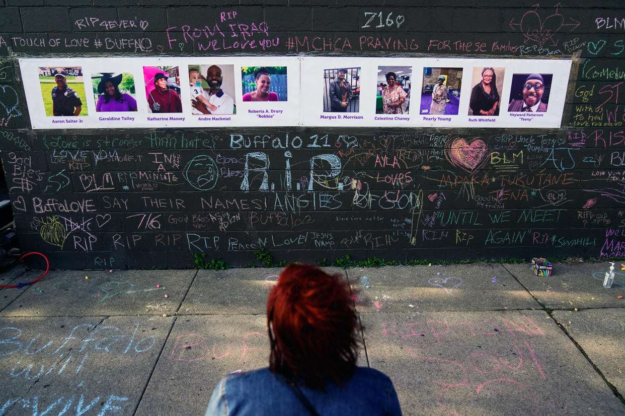 A person stands in front of a wall that holds pictures of the victims of a shooting with messages written in chalk around them.