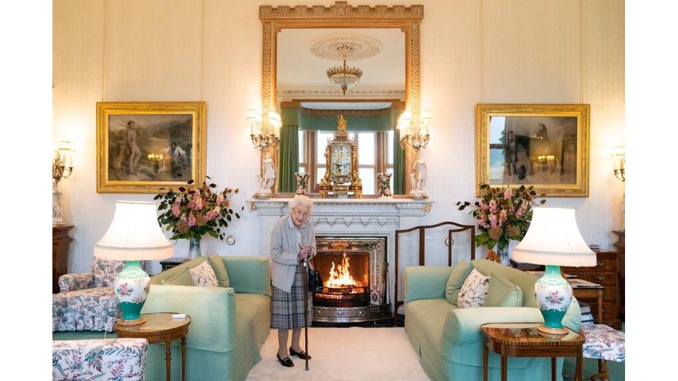The Queen standing in a light room with green sofas, floral lamps and a fireplace in the centre