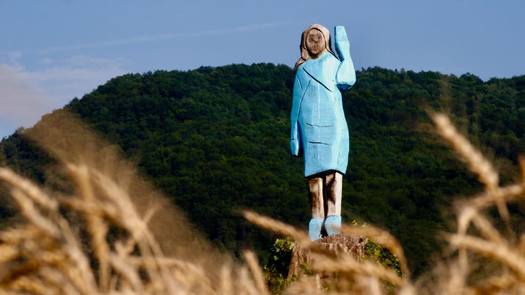 A crudely sculpted wooden sculpture of a woman wearing a light blue dress and waving with her left hand standing in a field with a green hill or mountain in the background