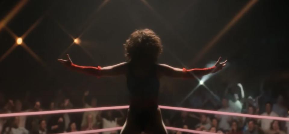 A female wrestler stands in the ring with her arms spread wide toward the crowd in a scene from 'GLOW' on Netflix