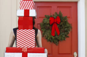 Woman holding stack of wrapped boxes, presents, or gifts. Presents cover her face as she stands in front of a red door with gree
