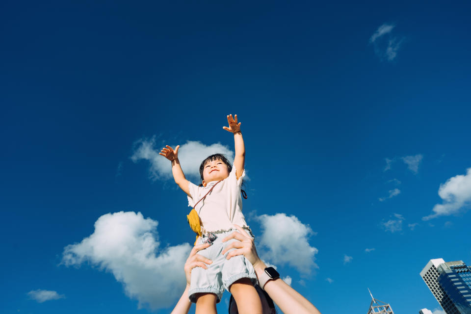 Happy Asian girl smiling joyfully and having fun outdoors while being carried by her mother and imagines flying in the air against beautiful blue sky in the city