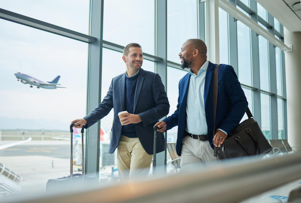 Two men walking in an airport terminal with carry-on luggage, a plane taking off outside the window