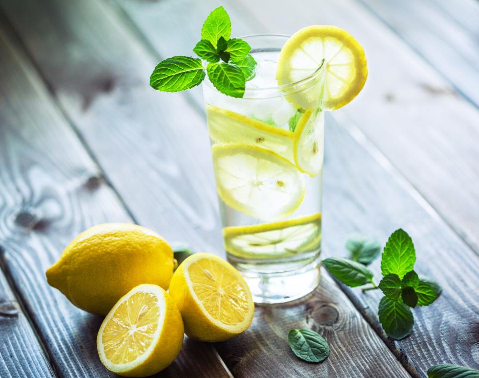 a glass of water with slices of lemon and a sprig of mint, next to more lemons and mint, all resting on a wooden table.