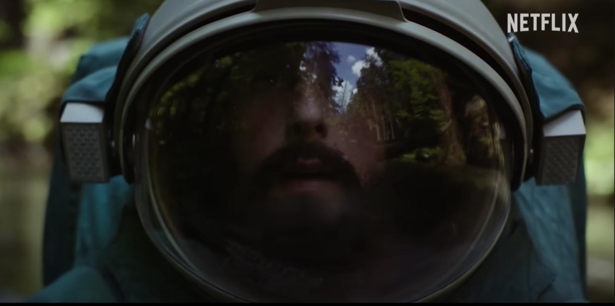  A bearded man in a spacesuit looks around confused in a forest. 
