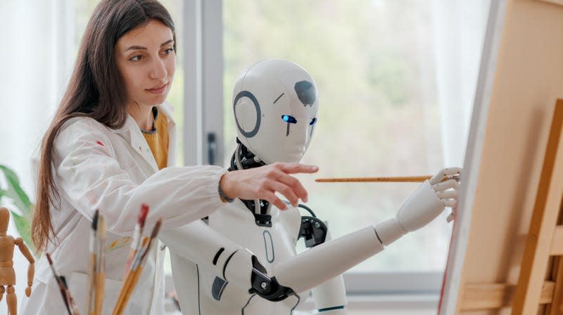 Artist woman teaching painting to a humanoid AI robot, she is pointing at canvas and giving advice to a robot surrounded by paint brushes and a canvas.
