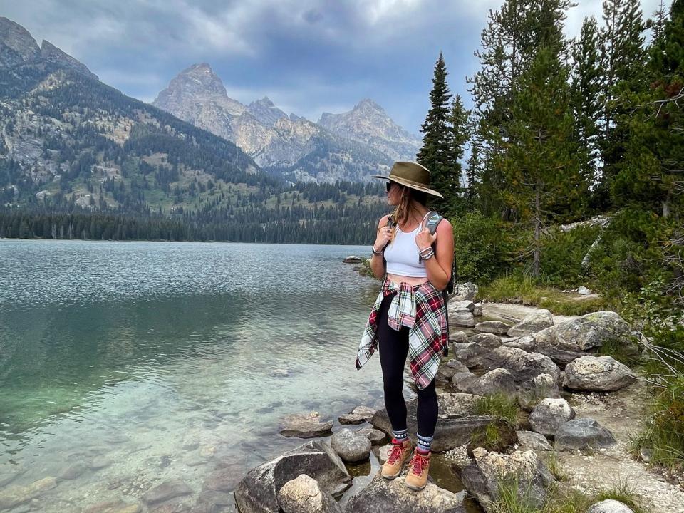 Emily stands on a rock and looks out at the green waters and large mountains at Grand Teton National Park. There are also trees behind her.