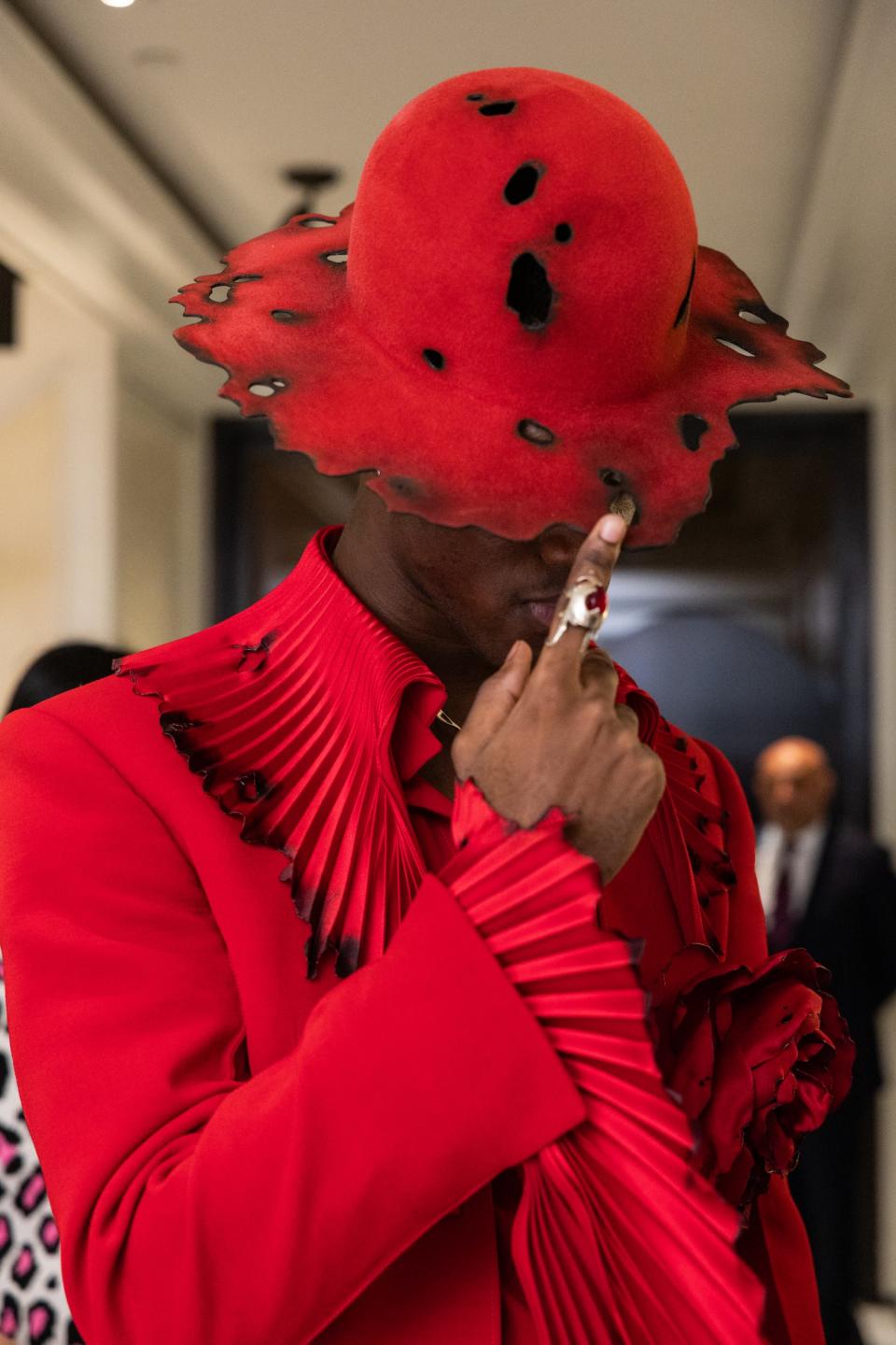 Individual in a red statement hat and fringed outfit posing with a finger on their lips