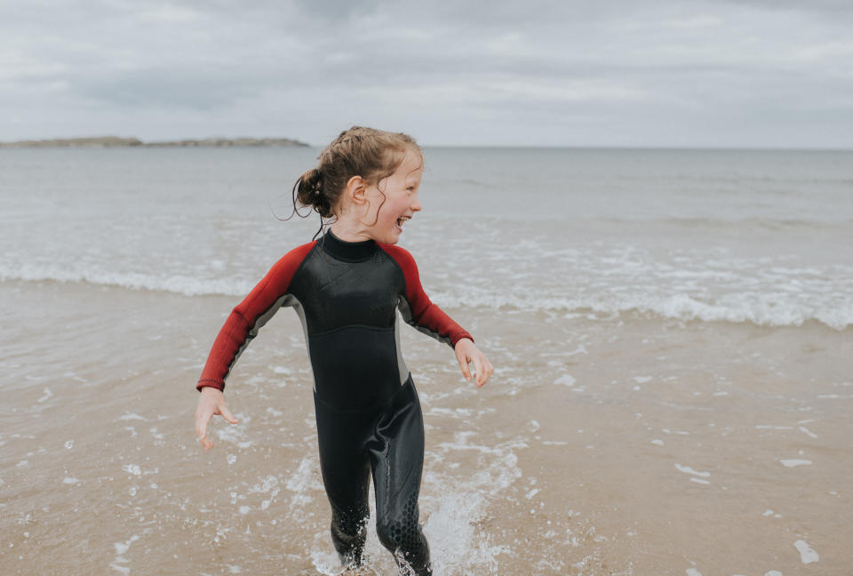 Joyful young child wearing a wetsuit paddles in the shallow tide. She looks away and laughs at something out of frame. Sky provides space for copy.