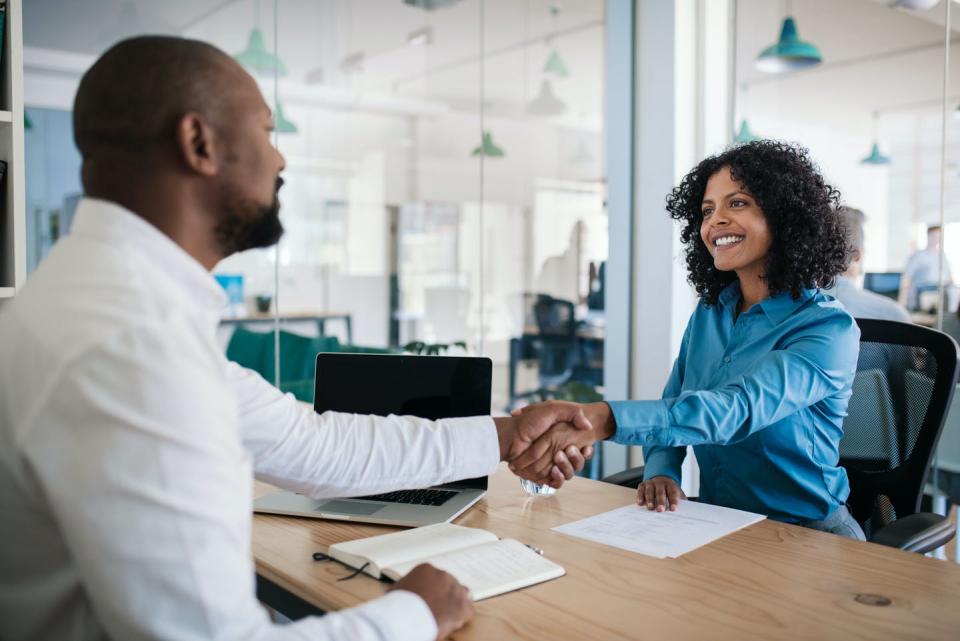 A man reaches his hand across a table to greet a woman, they're in an office setting, they're both smiling