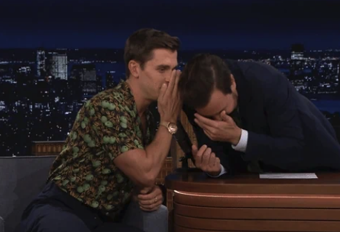 Antoni Porowski whispers to Jimmy Fallon on a late-night talk show, both laughing at the desk