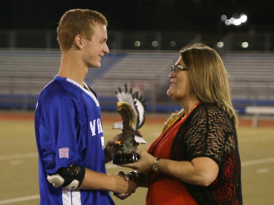 A white teenager in a blue lacrosse uniform accepts an eagle-shaped trophy from a woman in a red dress standing on a lacrosse field. Teenager is Daniel Penny, the person who put Jordan Neely in a chokehold on a Manhattan F train.