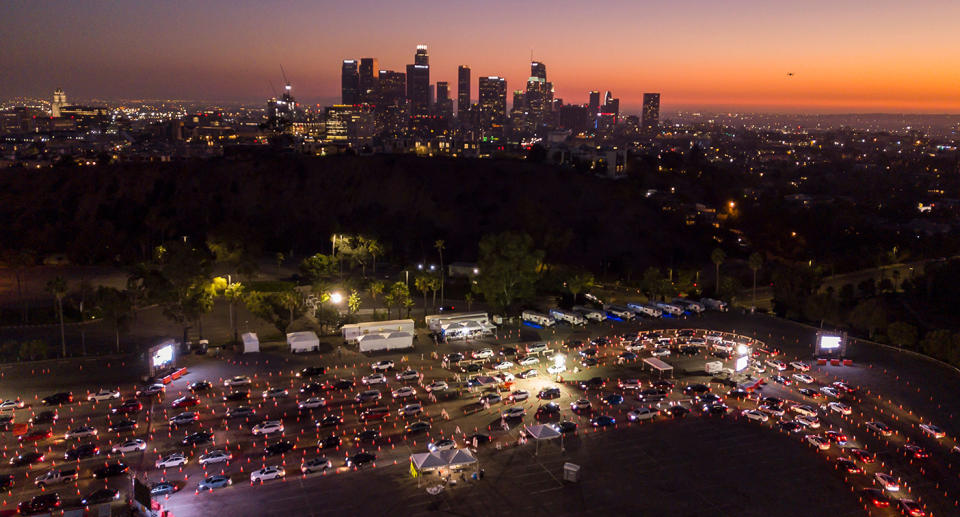 This aerial view shows cars lined up at the Dodger Stadium parking lot for Covid-19 testing with the downtown Los Angeles skyline in the distance.