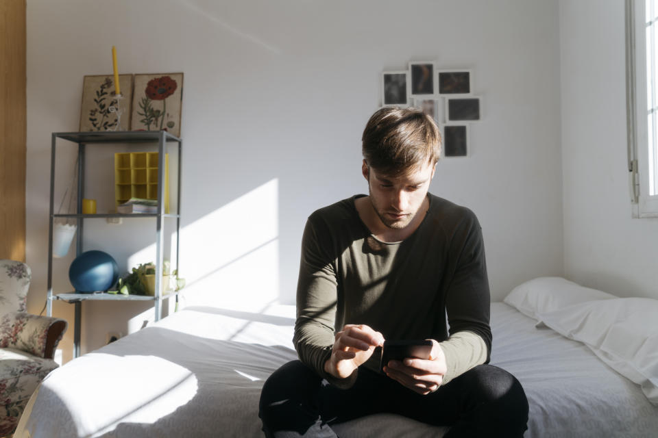 A man texting on his phone and sitting on his bed